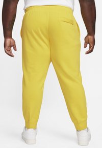 Pantalones de chándal amarillos con cinturilla elástica y bolsillos laterales. Piernas ajustadas con puños de canalé. Llevados con zapatillas blancas.