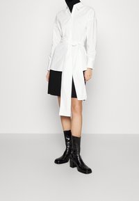 Femme portant une robe-chemise blanche ceinturée sur une robe noire, associée à des bottes en cuir noires à talons mi-mollet, debout sur un sol gris.