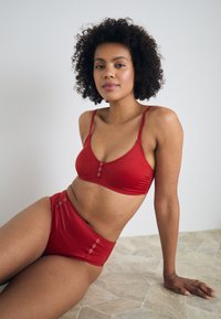 Crveni bikini set s braletom s okruglim dekolteom i prednjim gumbima, uparen s gaćicama visoko strukiranim koje imaju detalje s gumbima.
