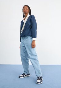 Marineblauwe lichtgewicht jas met witte kraag, lichtblauwe baggy broek en marineblauwe en witte sneakers, allemaal tegen een lichte achtergrond.