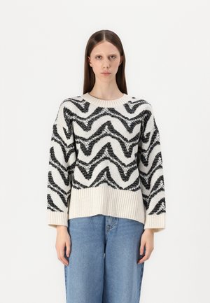 Maglione lavorato a maglia con una base crema e motivi ondulati neri, colletto e orlo a costine, e maniche ampie. Indossato con jeans blu.