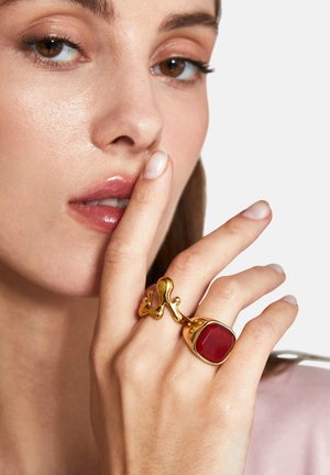 Goldring mit unregelmäßigem, organischem Design und einem roten Edelstein. Glatte metallische Oberfläche mit glänzender Textur, getragen an einer Hand.