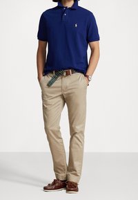 Námořnické modré polo tričko z bavlny s krátkými rukávy, zkombinované s světle béžovými chinos kalhotami a vícebarevným pruhovaným opaskem. Hnědé lodní boty dokončují look.