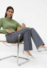 Maglione a coste verde chiaro, jeans grigi a zampa d'elefante e ballerine argentate con cinturino, seduta su una sedia moderna in cromo contro uno sfondo bianco.
