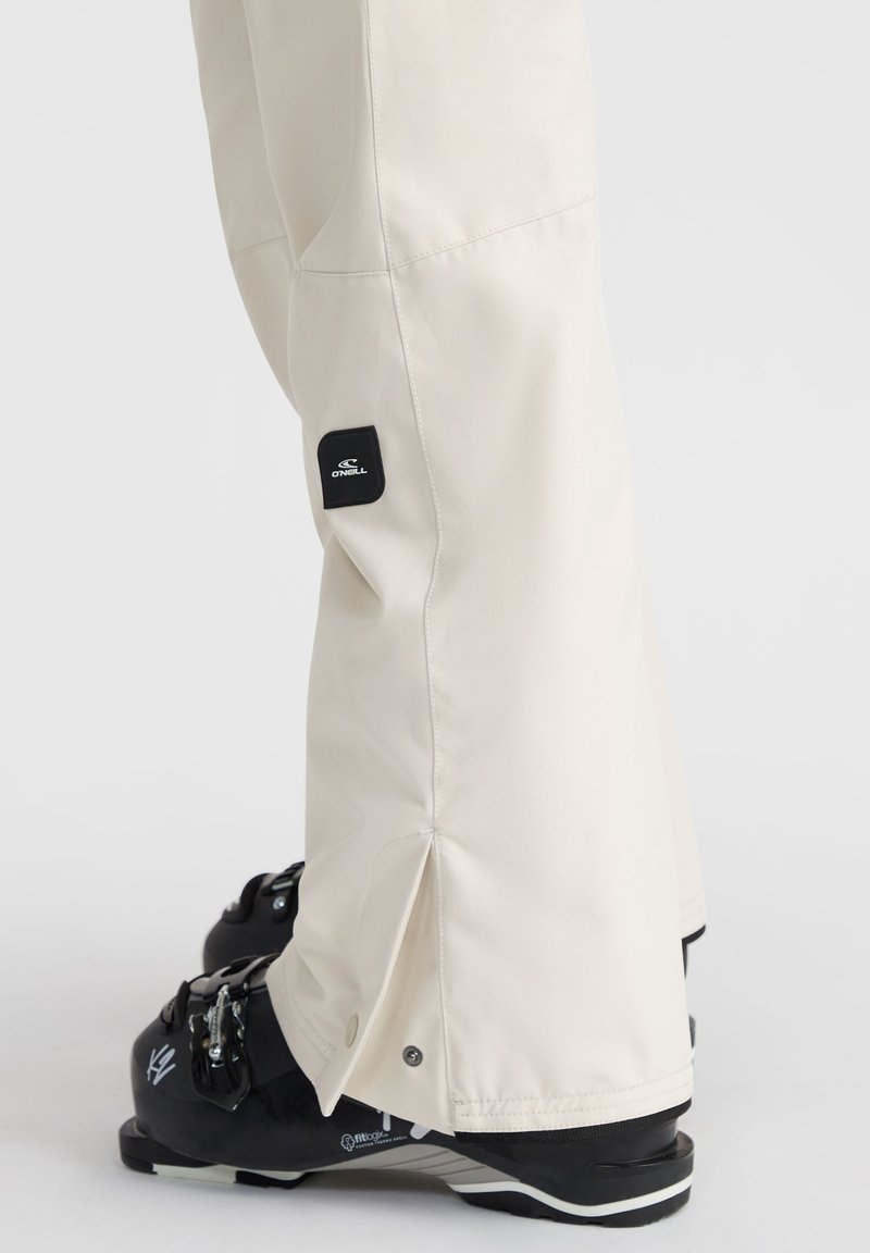 Pantalones de esquí beige con una textura suave, que presentan un parche de logo negro y cierres a presión en el dobladillo. Combinados con botas de esquí negras.