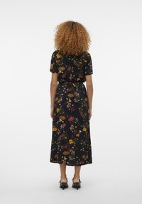 Kvinna med lockigt hår som bär en svart blommig midiklänning och svarta högklackade sandaler, stående vänd bort mot en enkel bakgrund.