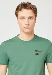 Groene katoenen T-shirt met ronde hals, korte mouwen en een geborduurd ontwerp "Vegetally Yours" met een groente-motief op de linkerborst.