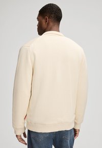 Veste tricotée de couleur crème avec des poignets et un ourlet côtelés, dotée d'un col et d'étiquettes sur les côtés, portée sur un jean en denim bleu.