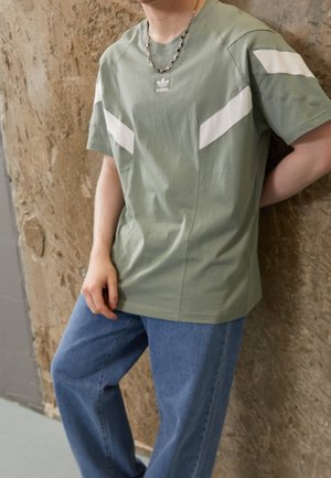 Zielona bawełniana koszulka z białymi ukośnymi paskami na ramionach i małym logo na piersi, w połączeniu z niebieskimi dżinsowymi spodniami.