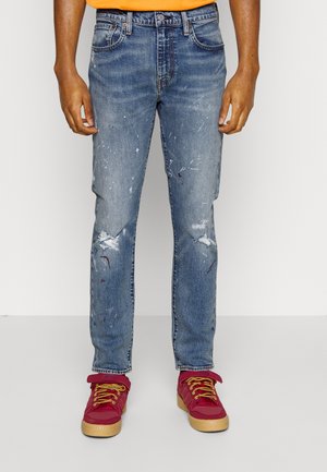 Hellblau beschädigte Jeans mit Farbspritzern, mit geradem Schnitt, fünf Taschen und mittlerer Leibhöhe; kombiniert mit roten Sneakers.