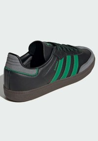 Black leather sneakers with three green stripes, grey accents, textured rubber sole, and patterned laces, featuring a stitched back detail.