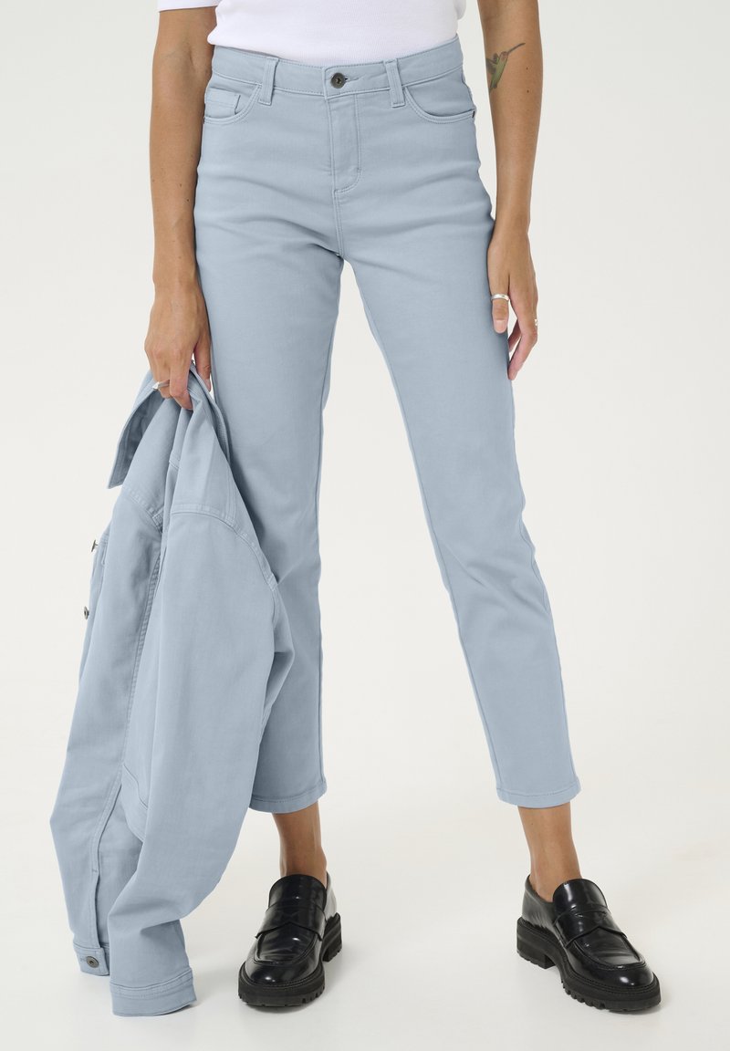 Jean bleu clair au design ajusté, taille haute, cinq poches et une longueur légèrement cropped, associé à une veste assortie et des mocassins noirs.