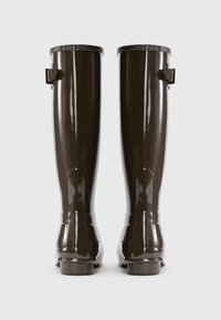 Botas de goma marrones, impermeables, con un acabado brillante, caña alta y detalle de correa ajustable en la parte superior. Suela plana y diseño de puntera redondeada.