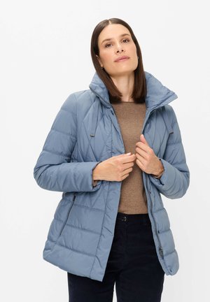Woman wearing a light blue quilted jacket over a brown sweater, adjusting the front zipper while standing on a plain white background.