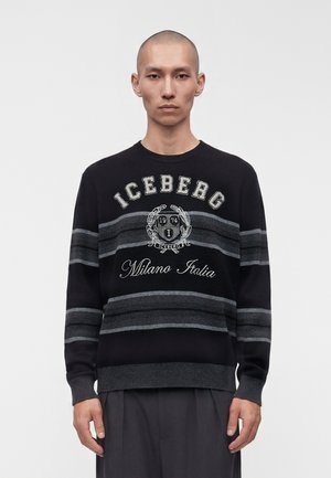 Mand med barberet hoved iført en sort og gråstribet sweater med teksten "ICEBERG Milano Italia" og emblem på ensfarvet baggrund.