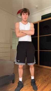 Bílý tílko, černé sportovní šortky s bílými pruhy a černé tenisky. Stojí s překříženýma rukama na dřevěné podlaze v místnosti.