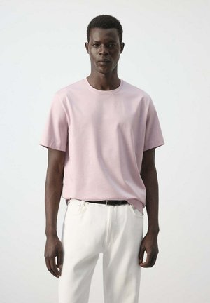 Homme portant un t-shirt rose clair à manches courtes et un pantalon blanc, debout devant un fond clair uni.