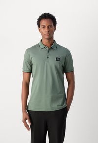 Polo shirt in saliegroen, gemaakt van zachte stof, met een klassieke kraag, een twee-knopen knoopsluiting en een klein zwart logo-patch op de borst.