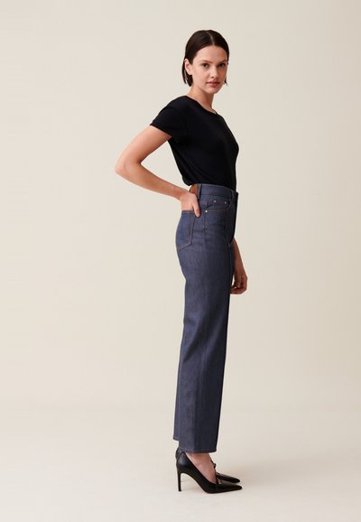 Jean en denim bleu marine taille haute avec une jambe droite, des poches latérales et des détails de couture subtils, associé à un t-shirt noir ajusté et des talons noirs.