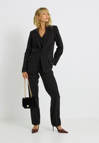 Costume noir sur mesure avec blazer structuré et pantalons droits. Accordé avec un sac à main en velours noir et des escarpins bordeaux pointus.