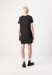 Person with short blonde hair stands facing away wearing a black short-sleeve dress, white socks, black chunky shoes, and hoop earrings.