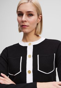 Black cardigan with white contrast trim, two front pockets, and gold buttons. Knitted texture with a fitted design.