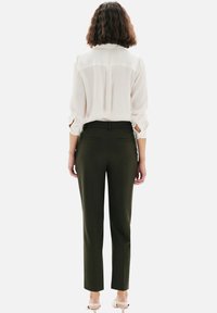 Blouse blanche à manches retroussées, associée à un pantalon taillé vert olive à coupe ajustée et poches arrière. Chaussures claires.