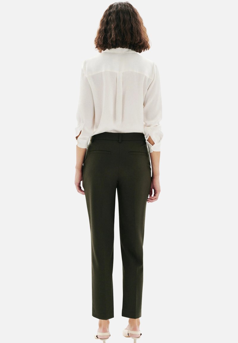 Blouse blanche à manches retroussées, associée à un pantalon taillé vert olive à coupe ajustée et poches arrière. Chaussures claires.