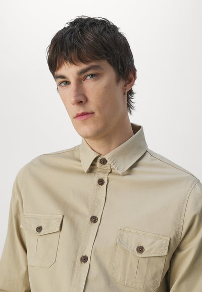 Beige button-up shirt made of cotton with two chest pockets. Features a classic collar and brown buttons, with a smooth texture.