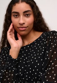 Blusa nera con pois bianchi, tessuto trasparente, polsini arricciati e maniche a sbuffo; il modello tocca il viso, sfondo neutro.