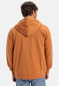 Orange hoodie med en dragsko på huvan, med en mjuk textur, ribbade manschetter och en avslappnad passform, bärs över blå denimjeans.