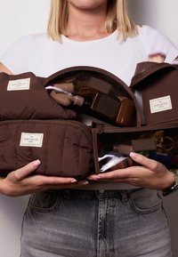 Brune, kviltede kosmetikposer med klare sektioner, der indeholder forskellige skønhedsprodukter. Har mærket "THE FLAT LAY CO. LONDON."