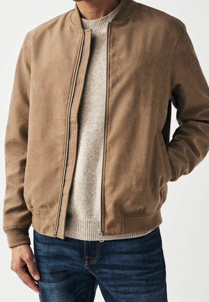 Mężczyzna w beżowej zamszowej kurtce bomberce, pod którą ma jasnoszary dziany sweter, i w ciemnoniebieskich dżinsach, z ręką w kieszeni kurtki.