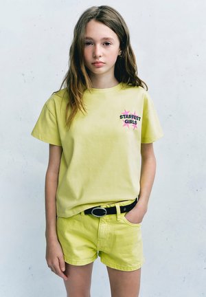 Fille aux longs cheveux bruns portant un t-shirt jaune clair Starfest Girls et un short assorti, debout avec une main dans la poche.