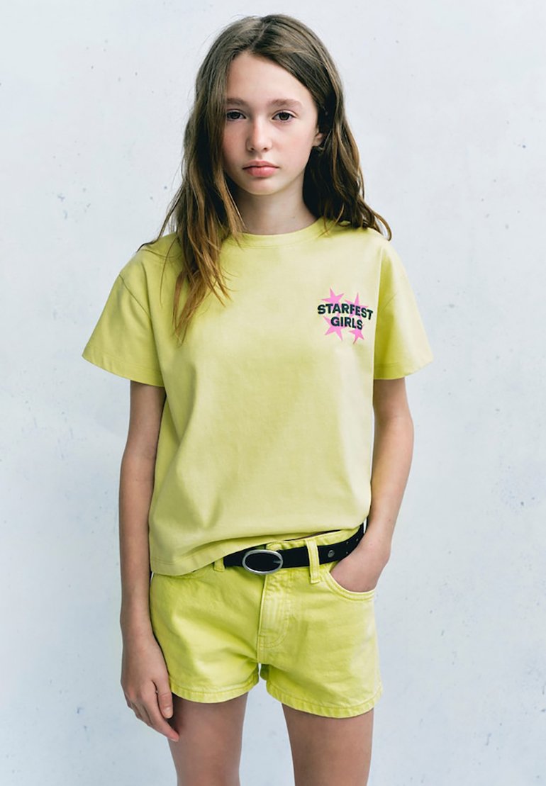 Fille aux longs cheveux bruns portant un t-shirt jaune clair Starfest Girls et un short assorti, debout avec une main dans la poche.