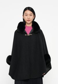 Cappotto di lana nera con colletto e polsini in pelliccia, caratterizzato da una chiusura dorata e accenti scuri, indossato sopra un maglione rosa e una gonna plissettata.