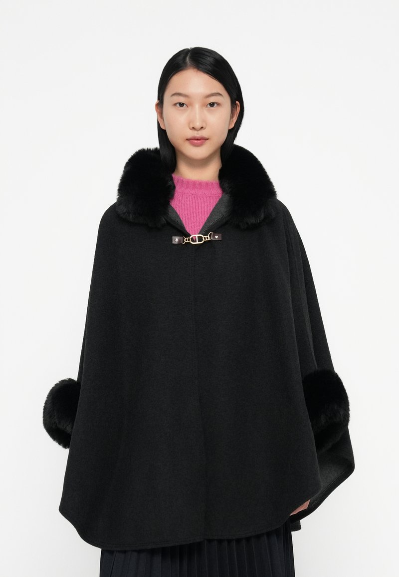 Cappotto di lana nera con colletto e polsini in pelliccia, caratterizzato da una chiusura dorata e accenti scuri, indossato sopra un maglione rosa e una gonna plissettata.