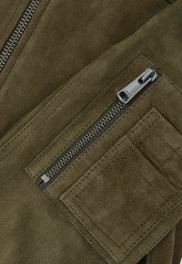 Õliva rohelise seemisnahast kangas, millel on tõmblukuga tasku ja hõbedased detailid. Silmatorkavad õmblused ja pehme tekstuur on esile toodud.