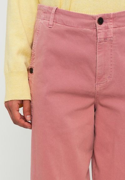 Person wearing high-waisted dusty pink pants with button and zipper closure, paired with a pale yellow long-sleeve sweater.