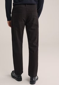Man in donkere bruine recht-toe-rechtdoor broek, zwarte leren schoenen en een donkerblauw overhemd met lange mouwen, staande tegen een effen achtergrond.