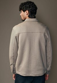 Beige struktureret jakke med knapkrave, synlige sømme og sideslidser. Bæres med mørke bukser, vist fra ryggen.