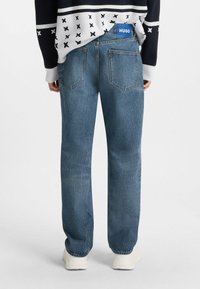 Jeans in denim blu con una vestibilità comoda, dotati di tasche posteriori e un'etichetta con logo blu sulla vita. Indossati con un maglione a fantasia.