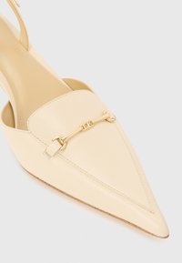 Beige Leder Slingback-Schuh mit spitzem Zehenbereich, goldenen Akzenten und Riemendetails. Glatte Textur und minimalistisches Design.