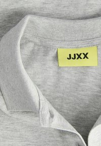 Grå polo skjorte med en ribbestrikket krage og knappestolpe. Merket er en lys gul med svart tekst "JJXX." Myk stofftekstur synlig.