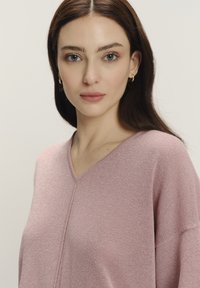 Bluzka z dekoltem w serek, z długim rękawem, w jasnoróżowym kolorze z delikatnym połyskiem, luźny krój, wykonana z miękkiej tkaniny, z wyraźnym detalem szwu na środku.
