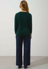 Pull en laine vert à coupe décontractée, manches trois-quarts ; associé à un pantalon en denim foncé à jambes larges ; texture lisse et design superposé.