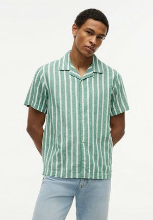 Homme portant une chemise à manches courtes boutonnée à rayures verticales vertes et blanches, avec un jean bleu clair, debout devant un fond blanc uni.