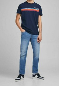 Mörkblå t-shirt med ett randigt mönster i röd, orange och blått på bröstet; kombinerad med ljusblå jeans och svarta sneakers.