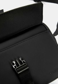 Borsa rettangolare nera con superficie liscia opaca, dotata di una tracolla nera regolabile e di una chiusura con un grande fermaglio di plastica nera.