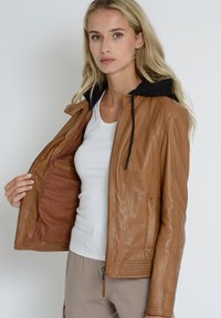 Veste en cuir marron avec capuche, dotée d'une fermeture éclair sur le devant, de coutures diagonales et d'une poche intérieure, portée sur un débardeur blanc.